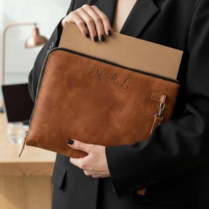 Puede incluir: Un portafolio de cuero marrón con cierre de cremallera y las iniciales "M.A.L." grabadas en la parte delantera. El portafolio está abierto, revelando un papel marrón en el interior. La persona lleva una blazer negra.