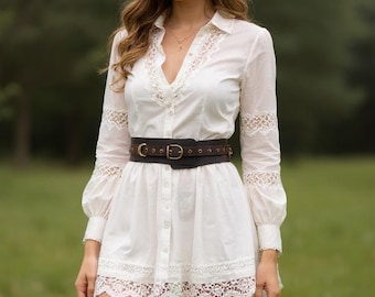 May include: A white dress with lace detailing, a brown belt, and black knee-high boots. The outfit is completed with a beige cowboy hat. The person is in a grassy field with trees in the background.