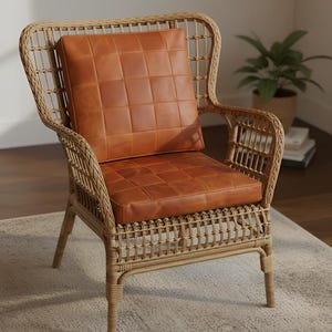 Op de afbeelding: Een rotan fauteuil met een hoge rugleuning en armleuningen. De zit- en rugkussens zijn gemaakt van bruin leer met een gewatteerd patroon. De stoel staat op een lichtgekleurd tapijt, met een plant in een pot zichtbaar op de achtergrond.