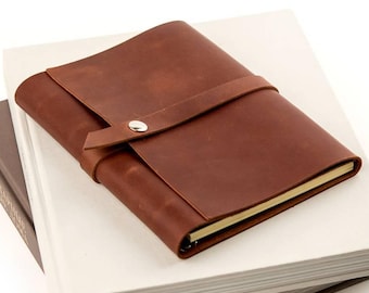 May include: A stack of three books, with a brown leather journal on top. The journal has a strap closure with a silver button. The books are a light gray, white, and dark gray.