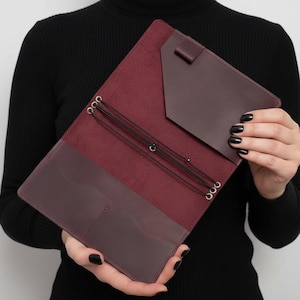 May include: Open burgundy leather journal with a matching pocket and elastic closures. The journal has a dark interior and is held open, showcasing the inside pockets and design. The leather is a deep, rich color.