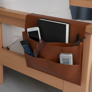 May include: A brown leather armchair organizer with multiple pockets. It holds a smartphone, glasses, a remote control, a notebook, a pen, and a small digital device. The organizer is attached to the arm of a wooden chair.