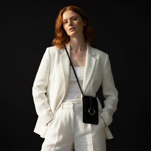 May include: A black crossbody phone bag with a silver ring detail. The bag is worn over a white outfit, including a blazer and pants. The background is a solid black.