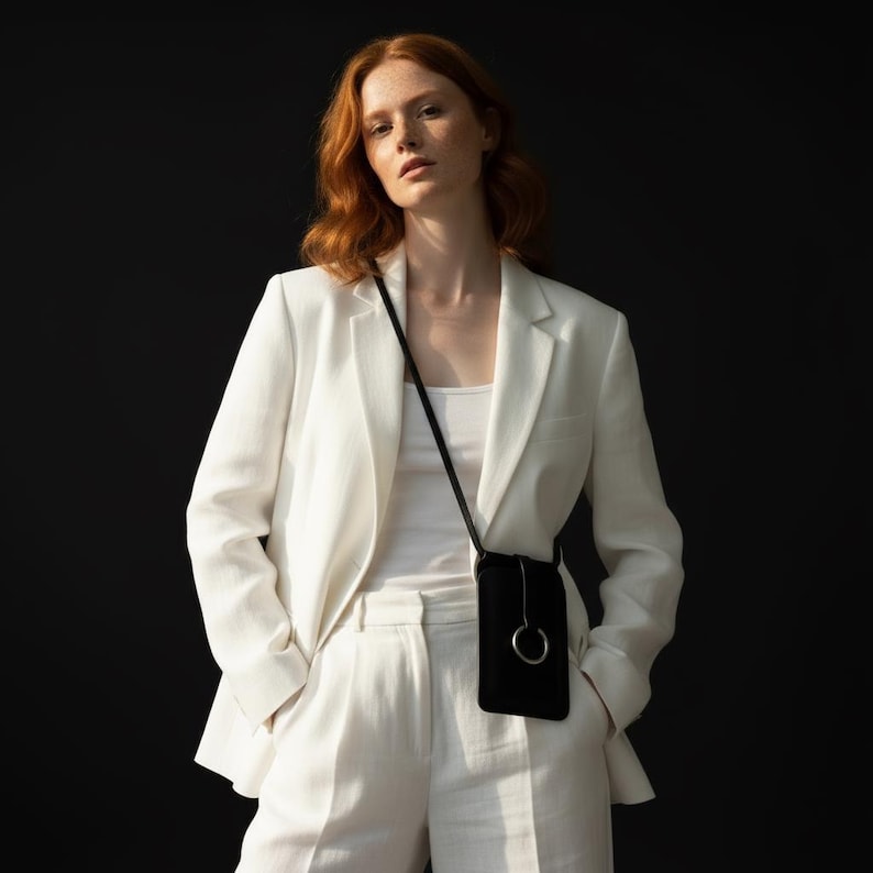 May include: A black crossbody phone bag with a silver ring detail. The bag is worn over a white outfit, including a blazer and pants. The background is a solid black.