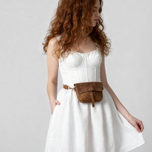 May include: A brown leather fanny pack with a buckle closure and a long strap. The fanny pack is worn around the waist with a brown leather belt. The model is wearing a white dress.