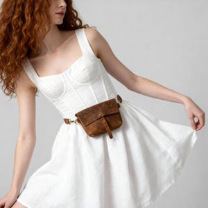 May include: A brown leather fanny pack with a buckle closure and a matching belt. The bag is worn around the waist over a white dress. The bag has a small embossed logo on the front.