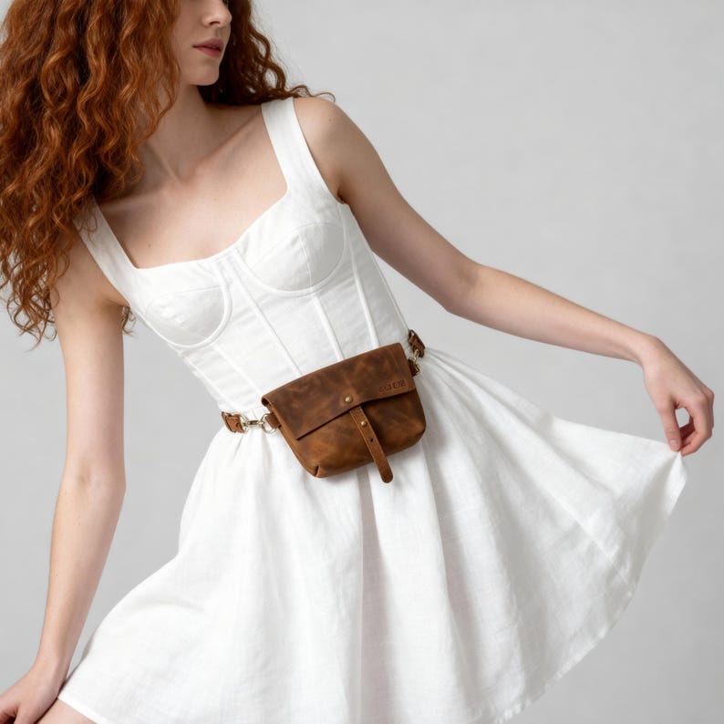 May include: A brown leather fanny pack with a buckle closure and a matching belt. The bag is worn around the waist over a white dress. The bag has a small embossed logo on the front.