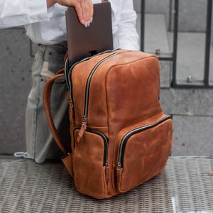 May include: A tan leather backpack featuring a large main compartment, a front zippered pocket, and side pockets. The bag has silver-toned metal zippers with leather pull tabs and adjustable shoulder straps, shown holding a dark gray laptop.