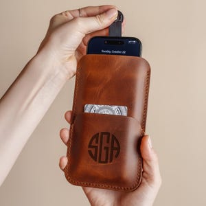 Könnte beinhalten: Eine braune Leder-Handyhülle mit Kartenfach und Monogramm. Die Hülle enthält ein Smartphone mit dem Datum "Sunday, October 22" auf dem Bildschirm. Eine Karte ist in der Vordertasche sichtbar.
