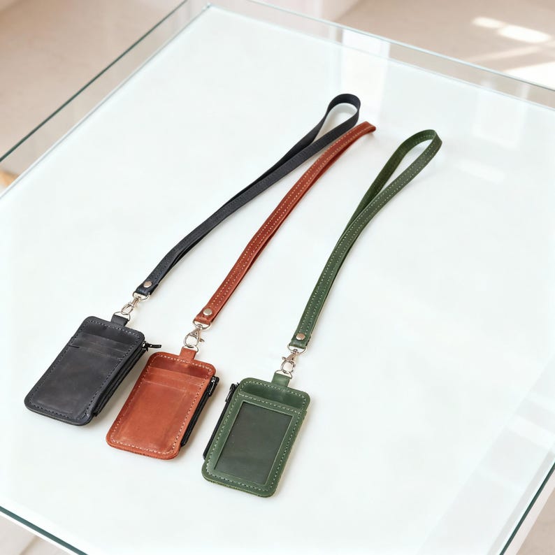 May include: Three leather ID badge holders in black, brown, and olive green, each with a matching lanyard. The holders have a clear window for displaying an ID and a zippered pocket. The lanyards are attached with silver-colored hardware.