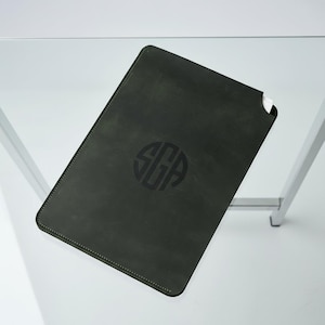 Könnte beinhalten: Dunkelgrüne Tablet-Hülle aus Leder mit einer genähten Bordüre und einem kreisförmigen Monogramm in der Mitte. Das Monogramm zeigt die Buchstaben "SGA". Die Hülle ist rechteckig und scheint aus weichem, geschmeidigem Material zu sein.