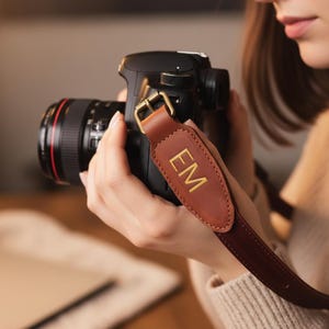 May include: A black DSLR camera with a brown leather strap. The strap has gold-colored hardware and the initials "EM" in gold lettering. The camera lens is black with a red accent.