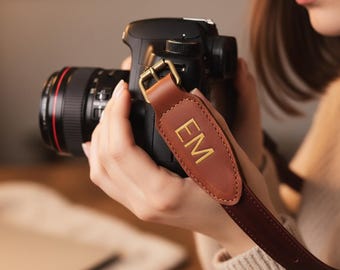 May include: A black DSLR camera with a brown leather strap. The strap has gold-colored hardware and the initials "EM" in gold lettering. The camera lens is black with a red accent.