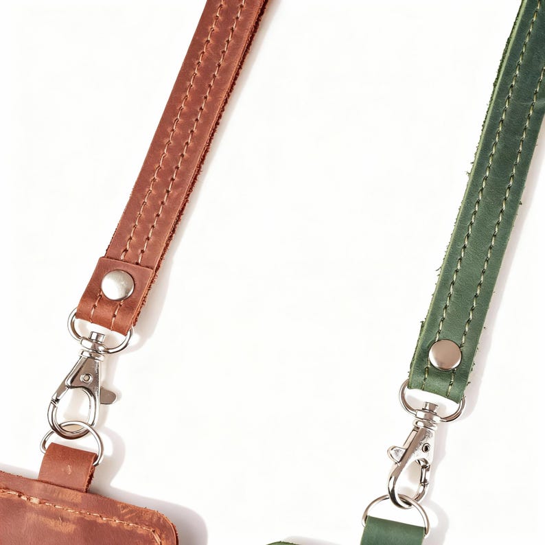 May include: Close-up of two leather straps with metal clasps. One strap is brown, the other is green. Both straps have stitched edges and silver-colored metal hardware. The straps are likely for a bag or other accessory.