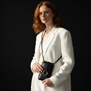May include: A black leather crossbody phone bag with a silver ring detail. The bag is worn over the shoulder. The person is wearing a white blazer and top. The background is black.