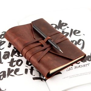 May include: A brown leather journal with a pencil secured by a leather strap. The journal is open on a page with black text. The journal is a rich, warm brown color.