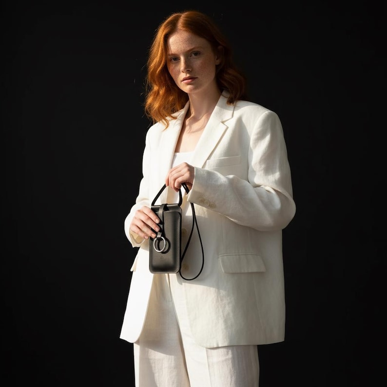 May include: A black leather phone crossbody bag with a silver ring detail. The bag has a long black strap. The person in the image is wearing a white blazer and pants. The background is black.