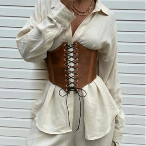 May include: A brown corset with black lacing is worn over a cream-colored shirt. The corset has a structured design and is laced up the front. The shirt has long sleeves and a button-down style. The background is a white wall.