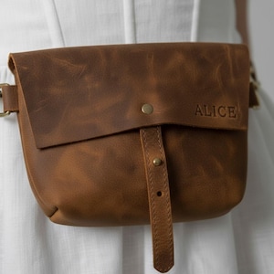 May include: A brown leather fanny pack with the name "ALICE" embossed on the front. The bag features a flap closure with a brass button and a matching adjustable strap. The bag is worn around the waist.