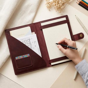 Puede incluir: Una funda de cuero color burdeos oscuro para cuaderno con cierre de broche, un bolsillo interior con un cuaderno y un documento, y un bloc de notas cuadriculado sujeto por dos correas horizontales. Un bolígrafo negro se sostiene sobre el bloc.
