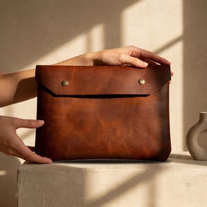 Puede incluir: Una funda para portátil de cuero marrón con cierre de solapa asegurado por dos botones de presión de color latón. La funda tiene una superficie texturizada y está sujeta por dos manos.