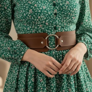 Könnte beinhalten: Ein brauner Ledergürtel mit einer großen silberfarbenen kreisförmigen Schnalle. Der Gürtel wird über einem grünen Kleid mit Blumenmuster getragen. Der Gürtel hat ein breites Design und wird mit einer Metallschnalle befestigt.