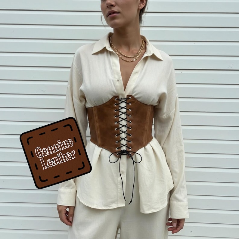 May include: A brown genuine leather corset with a lace-up front. The corset is worn over a cream-colored button-down shirt and pants. The image includes a square sign that says "Genuine Leather".
