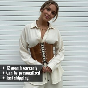 May include: A brown corset with black lacing is worn over a cream-colored button-down shirt. The corset is the focal point, with a structured design and visible lacing. The image also includes the text: "12 month warranty", "Can be personalized", and "Fast shipping".