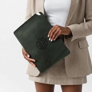 May include: Dark green leather laptop sleeve with a monogram design. The sleeve is held by a person wearing a tan blazer and skirt. The monogram is in a darker shade of green.