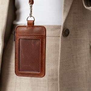 May include: A brown leather ID badge holder with a clear window and a matching leather lanyard. The holder is rectangular with stitched edges and a silver-colored clasp. The background includes a beige blazer.