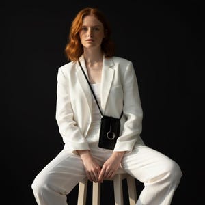 May include: A black crossbody phone bag with a silver ring detail. The bag is worn over a white top and a white blazer. The person is sitting on a wooden stool. The background is black.