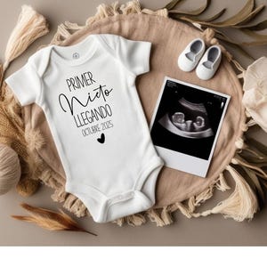 May include: White baby onesie with black text that reads "Primer Nido Llegando Octubre 2023" with a small black heart. A black and white ultrasound image is in the background.