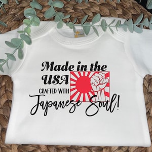 May include: White baby bodysuit with the text "Made in the USA Crafted with Japanese Soul!" in black script. The design includes a red sun with white rays and a clenched fist.