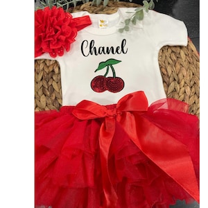May include: A white baby bodysuit with the name "Chanel" in black script and a cherry design. The outfit includes a red tutu skirt with a large bow and a matching red flower headband. The ensemble is displayed on a woven surface.