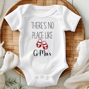 Puede incluir: Un body blanco para bebé con texto negro que dice "There's no place like G-Ma's" con un par de zapatos rojos con corazones blancos.