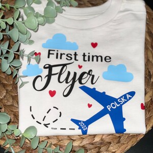 May include: White t-shirt with a blue airplane graphic and the text "First time Flyer" in black. The airplane has the word "POLSKA" written on it. The year 2022 is written on the airplane's wing.