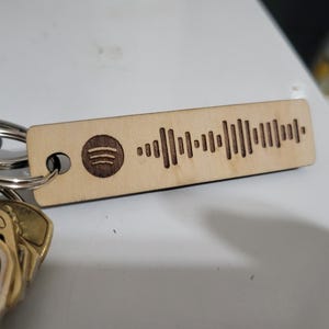 May include: A rectangular light-coloured wooden keyring featuring a dark brown engraved Spotify logo and a corresponding sound wave pattern. The keyring is attached to a metal ring that holds several keys with blue and gold-toned metal heads.