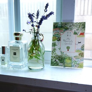 May include: Two clear glass bottles of perfume with silver caps and labels. The bottle on the left is smaller and the bottle on the right is larger. The larger bottle has a label that reads "Jo Malone London" and "English Pear & Freesia". A bouquet of lavender flowers in a clear glass vase is in the center of the image. A card with the text "My Heavenly Garden" is on the right side of the image.