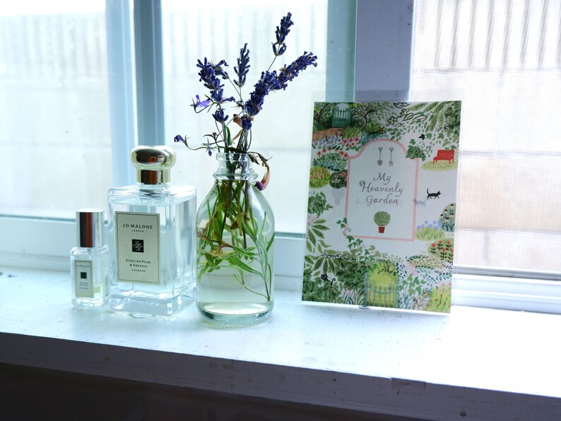 May include: Two clear glass bottles of perfume with silver caps and labels. The bottle on the left is smaller and the bottle on the right is larger. The larger bottle has a label that reads "Jo Malone London" and "English Pear & Freesia". A bouquet of lavender flowers in a clear glass vase is in the center of the image. A card with the text "My Heavenly Garden" is on the right side of the image.