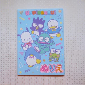 May include: A blue notebook with a light blue background featuring a group of cartoon characters from the Japanese children's television show "Kero Kero Keroppi". The characters include Keroppi, the frog, Tuxedo Sam, the penguin, Pikki, the dog, and a few others. The notebook has the text "Hapidanbui" and "めりす" printed on it.