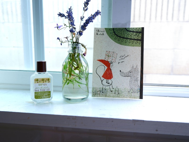May include: A clear glass bottle with a brown lid filled with lavender flowers and green stems. A small bottle of tea tree oil is next to the vase. A book with the title "Shimai Koibito" is on the windowsill behind the bottles.