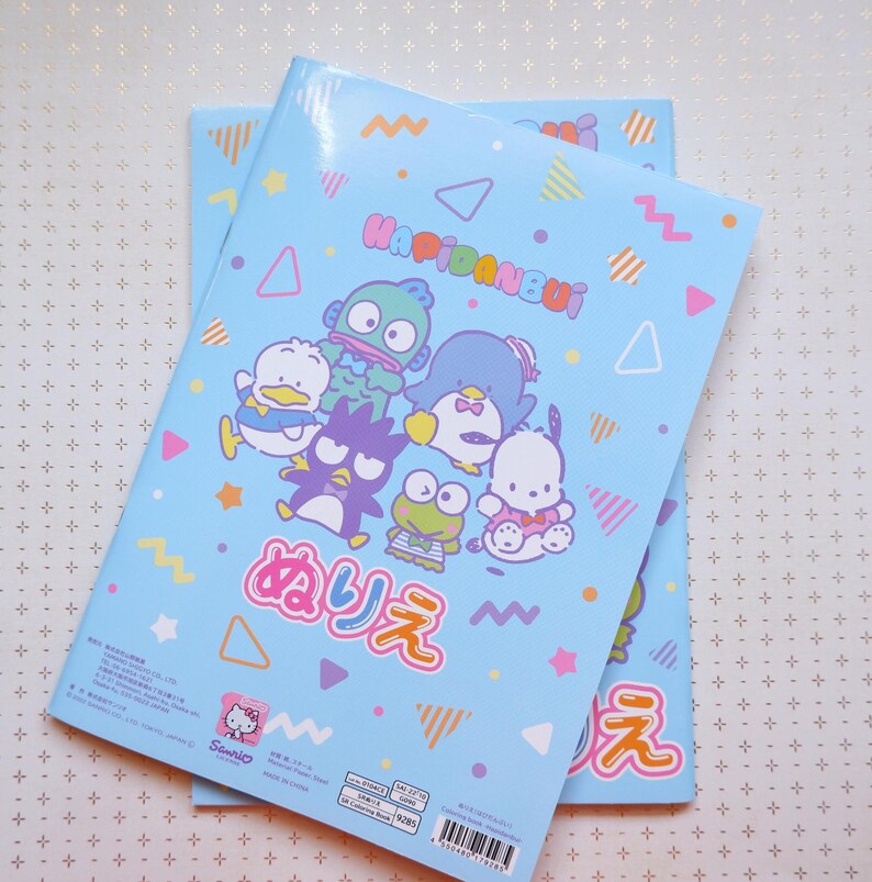 May include: Two blue notebooks with colorful geometric patterns and cartoon characters. The front cover features a group of cartoon characters including a penguin, a frog, a duck, and a monster. The text on the cover reads "Happidanbui" and "みりえ".
