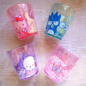 May include: Four clear plastic cups with colorful Sanrio character designs. Each cup features a different character, including Hello Kitty, Keroppi, and Cinnamoroll. The cups have a pink, red, blue, and purple color scheme.
