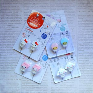 May include: Four packages of Hello Kitty and My Melody cable protectors for iPhone. Each package contains two cable protectors, one pink and one blue. The packages are labeled with the Hello Kitty and My Melody characters and the text "Cable Protector".