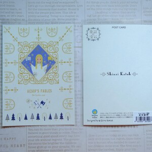May include: A postcard featuring a whimsical illustration of a woman in a blue dress with a golden crown, surrounded by a decorative frame. The text "Aesop's Fables" is printed in gold lettering. The back of the postcard features the name "Shinzi Katoh" in black lettering.