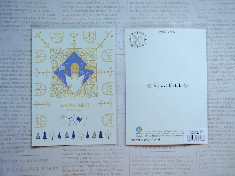 May include: A postcard featuring a whimsical illustration of a woman in a blue dress with a golden crown, surrounded by a decorative frame. The text "Aesop's Fables" is printed in gold lettering. The back of the postcard features the name "Shinzi Katoh" in black lettering.