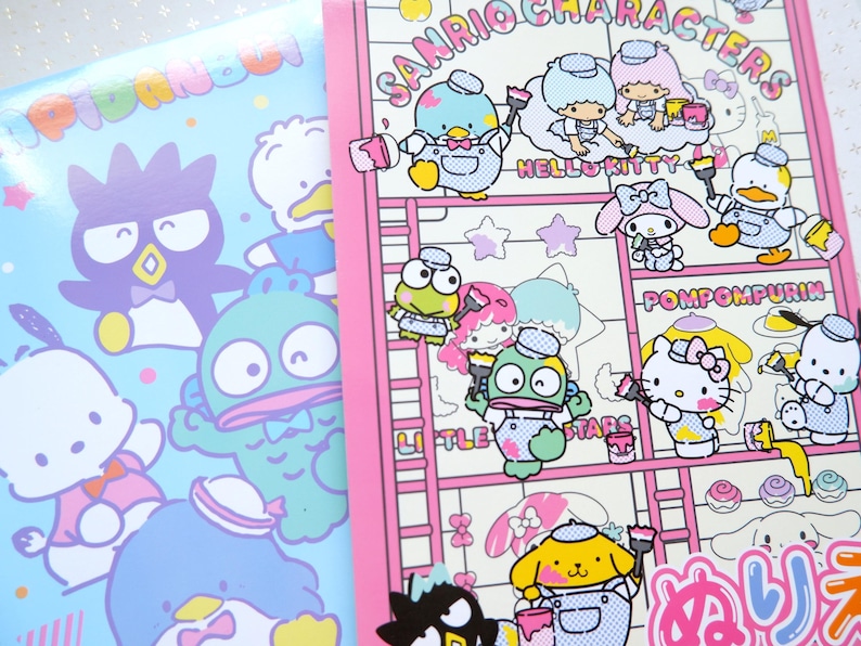 May include: A pink and white notebook with a Sanrio character theme. The cover features Hello Kitty, Pompompurin, and other Sanrio characters painting on a ladder. The text "Sanrio Characters" is printed in black on the cover.