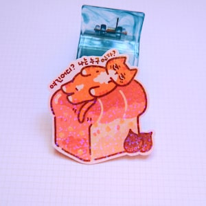 May include: A sticker featuring a cartoon orange cat sleeping on a loaf of bread with a glittery finish. The sticker has text in Korean. A clear blue object is in the background.