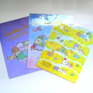 May include: Three Sumikkogurashi-themed file folders in purple, blue, and yellow. Each folder features a different design with various Sumikkogurashi characters. The purple folder has the text "Sumikkogurashi" in gold script. The folders are made of a clear plastic material.