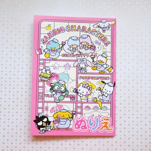 May include: A pink notebook with a colorful illustration of Sanrio characters painting. The characters include Hello Kitty, Pompompurin, Little Twin Stars, and others. The notebook has the text "Sanrio Characters" and "メルヘン" in Japanese.